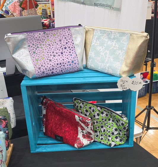 A craft fair booth displaying various fabric pouches and zippered bags in different colors and patterns, arranged on a black tablecloth next to a bright blue wooden crate.
